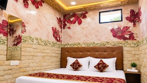 Een slaapkamer met een bakstenen muur, een groot bed met witte beddengoed en rode bloemenkussens, een houten hoofdbord, een klein nachtkastje met een plant, en een spiegel aan de muur.
