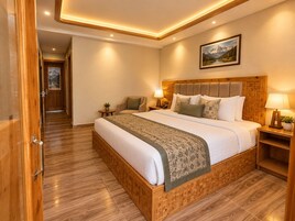 A hotel room with a wooden bed frame, a large bed, a nightstand with a lamp, a wall-mounted painting, and a wooden floor.