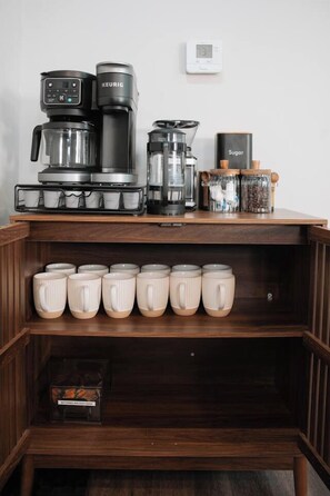 Ein Kaffee-Bereich mit einer Keurig-Kaffeemaschine, einem Mahlgerät und Zuckerbehältern auf einem Holzregal.