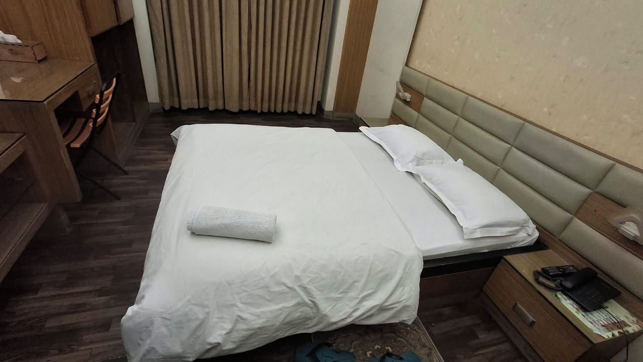 A hotel room with a bed, a wooden desk, a chair, and a bedside table with a phone.