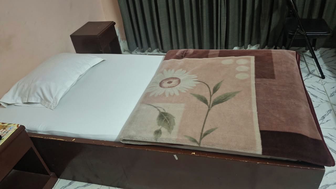 Una cama con sábana blanca y manta estampada, una mesita de noche de madera, un aparador de color oscuro y una silla en una habitación con cortinas.