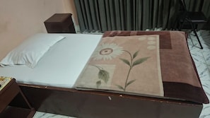 Un lit avec un drap blanc et une couverture à motifs floraux, une petite table de chevet en bois, une commode de couleur sombre et une chaise dans une pièce avec des rideaux.