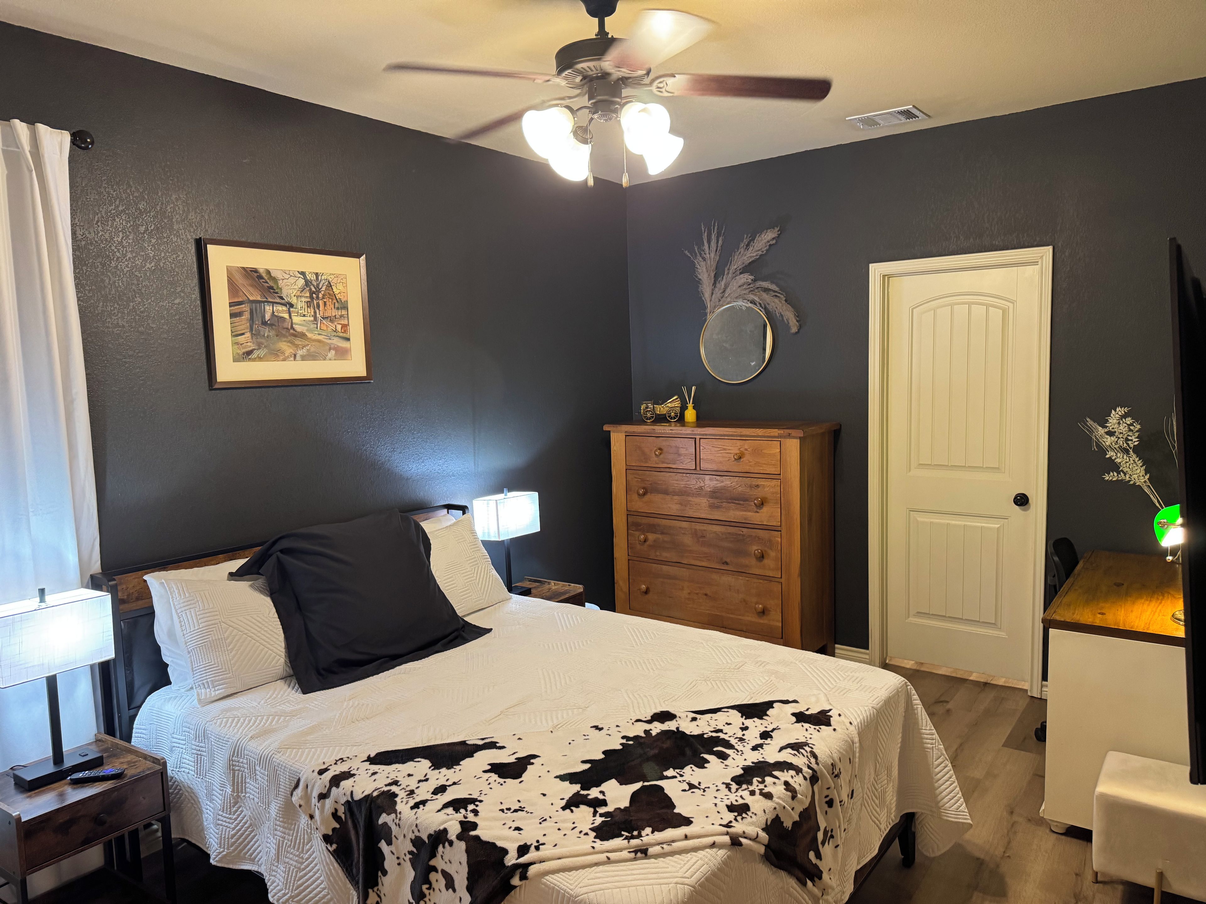 Une chambre à coucher comprenant un lit, des tables de chevet, une commode, un ventilateur de plafond, une porte et un miroir fixé au mur.