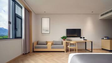 A modern hotel room with a bed, a desk, a sofa, a TV, and a window with a view of mountains.