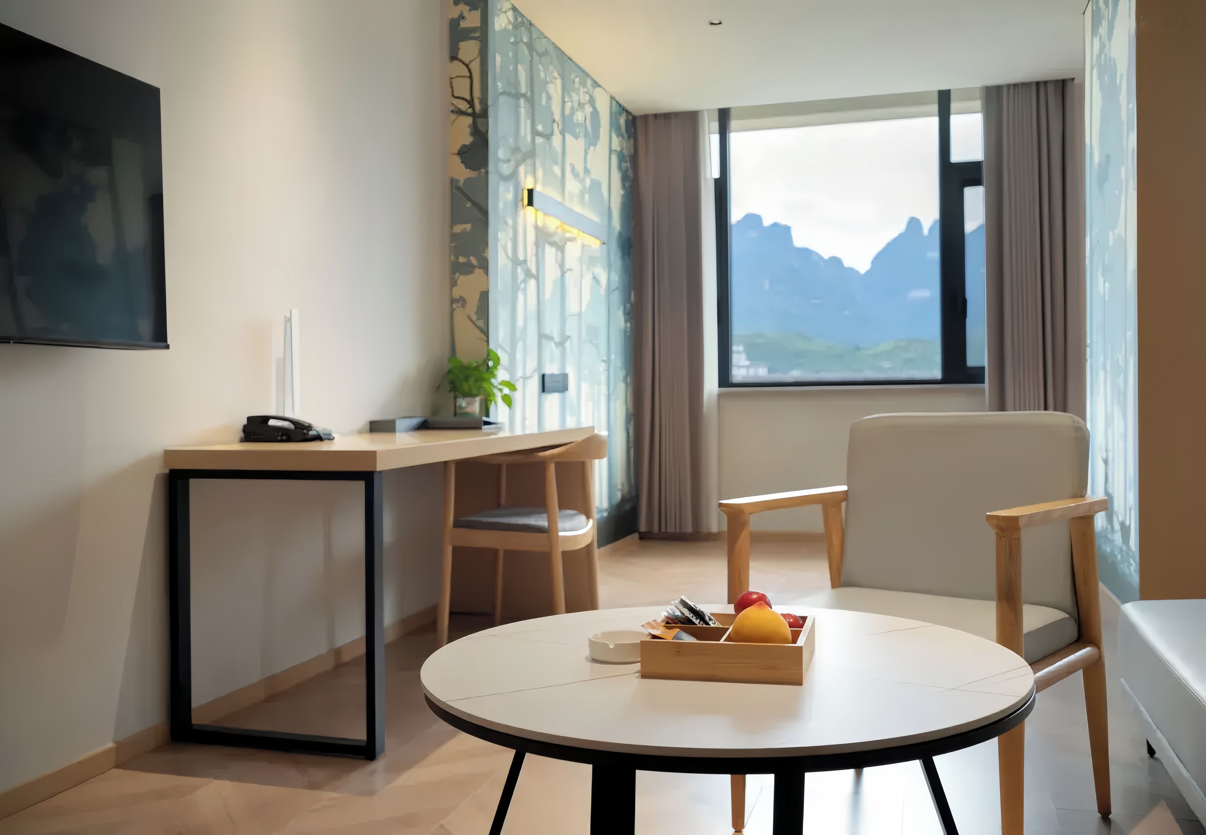 Una habitación de hotel moderna con una mesa redonda, dos sillas, un escritorio, un teléfono, una planta y una ventana con vistas a la montaña.