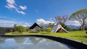 A pool with a clear reflection, a modern building, and a tent in a grassy area with trees and mountains in the background.