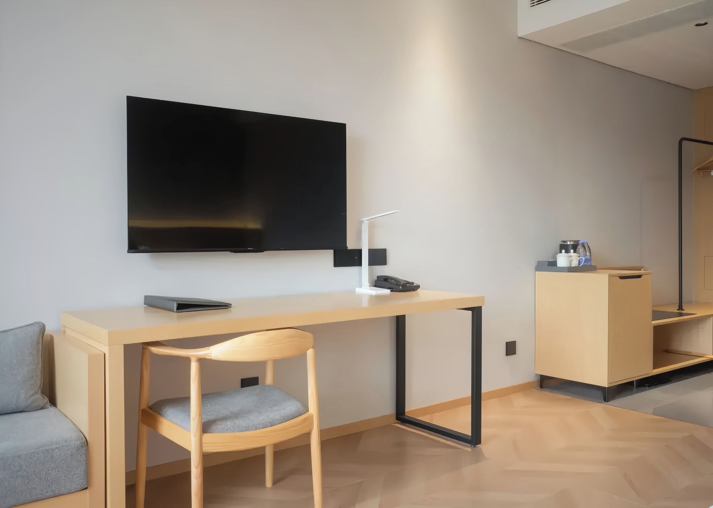 A modern, minimalist room with a flat-screen TV mounted on the wall, a wooden desk with a chair, and a small cabinet with a kettle.