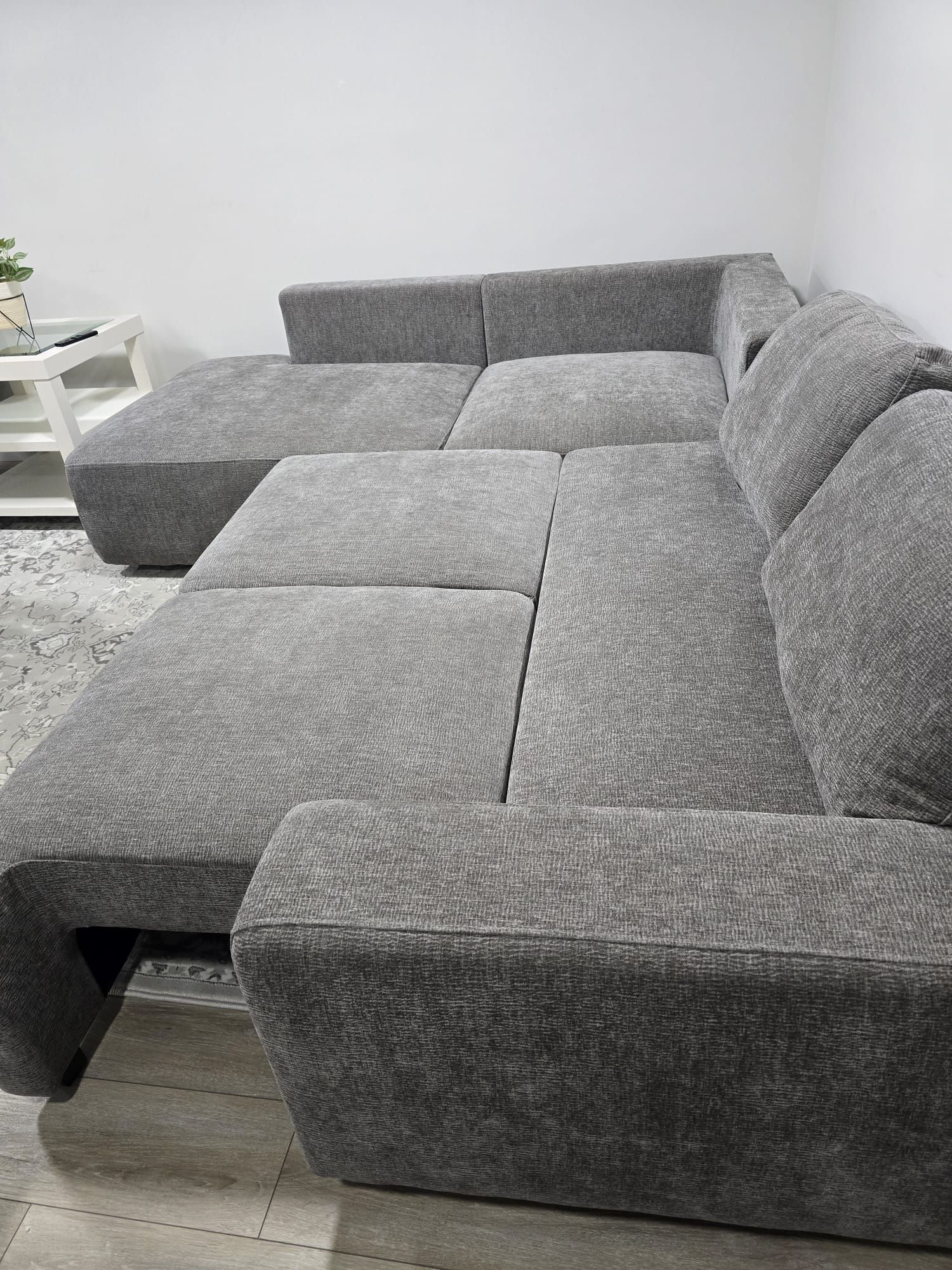 A grey sectional sofa with a matching ottoman in a room with a white wall and a small white side table.