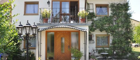 Una casa de dos pisos con puerta principal de madera, un balcón con plantas en macetas y un pequeño jardín con mesa y sillas.