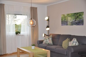 A living room with a grey sofa, a wooden coffee table, a hanging light fixture, and a painting on the wall. - Vacation Rental: 78 square meters, 1 separate room Bedroom, outdoor seating area, patio (Burghausen)
