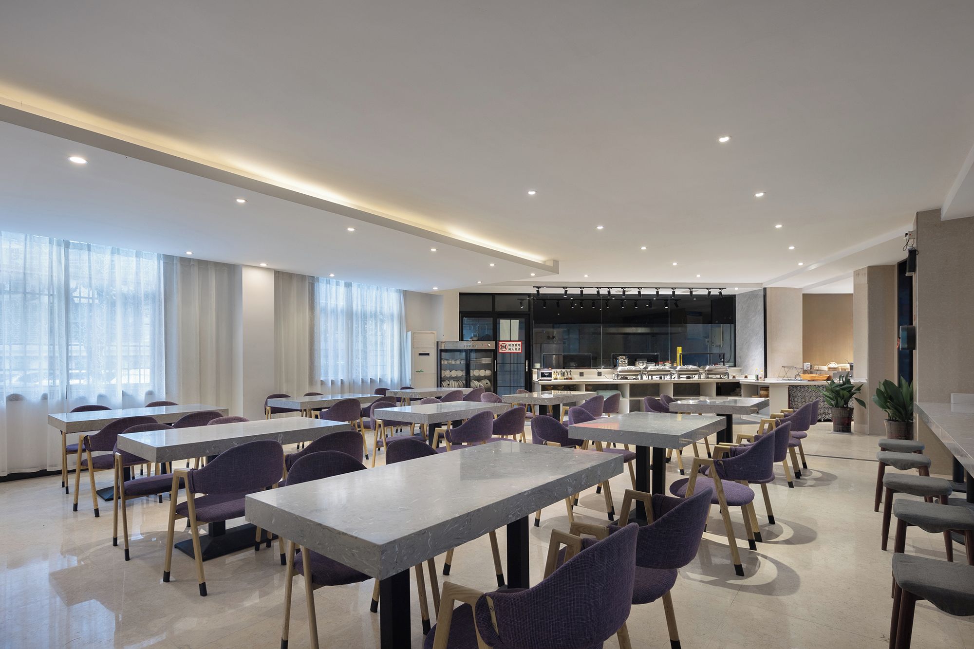 A modern dining area with multiple tables and chairs, a kitchen area with appliances, and a large screen for presentations.