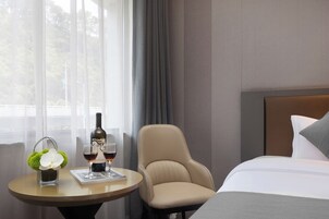 A hotel room with a bed, a round table with a bottle and glasses, a chair, and a window with curtains.