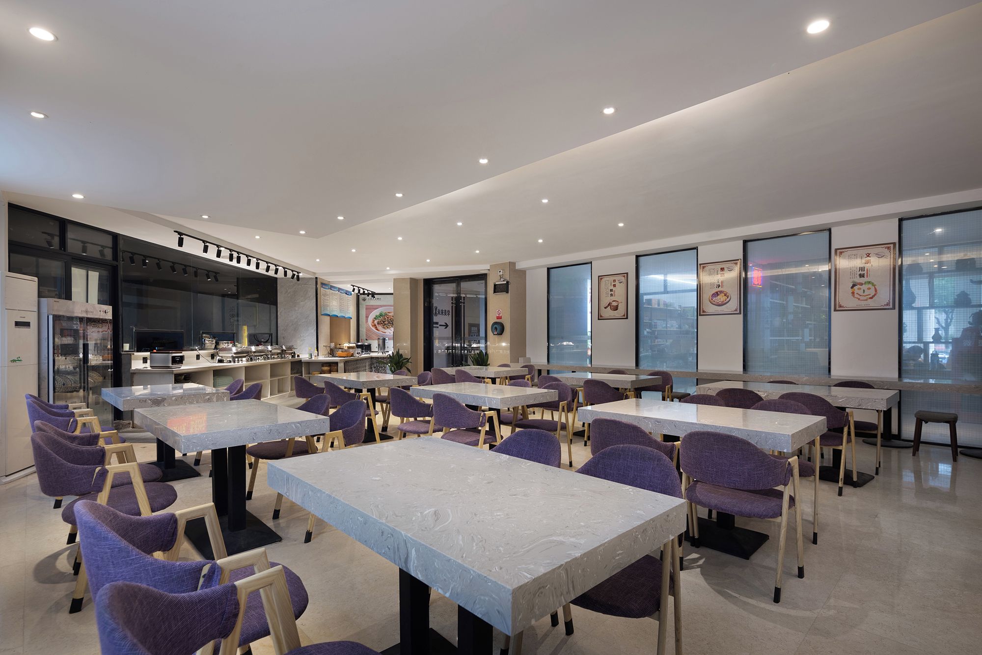 A modern dining area with multiple tables and chairs, a kitchen in the background, and large windows.