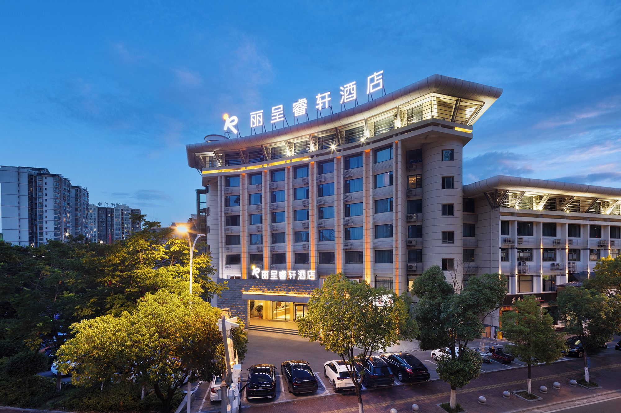 A multi-story hotel with a prominent sign at the top, a parking lot with several cars, and surrounding greenery.