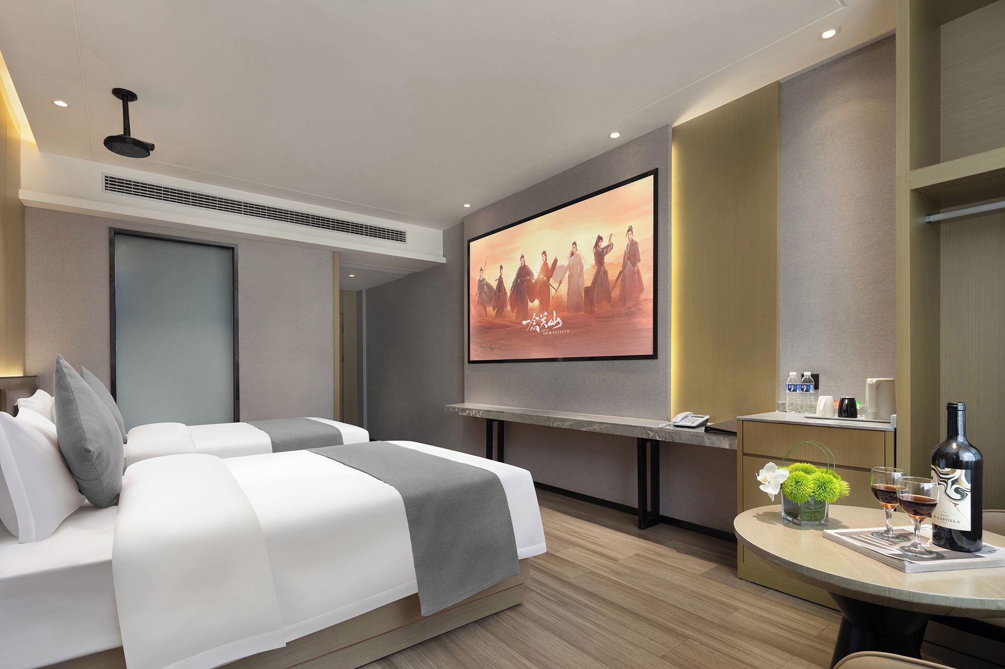 A modern hotel room with a bed, a TV screen, a desk with a bottle of wine, and a vase with flowers.