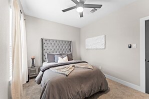 A bedroom with a tufted headboard, a bed with a brown comforter, a bedside table with a vase, and a ceiling fan. - Entire duplex | Two homes, one stay (Houston)