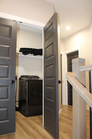 A laundry room with a washing machine and dryer, a door leading to a hallway, and a staircase with a wooden handrail. - Entire duplex | Two homes, one stay (Houston)