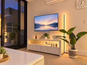 A modern living room with a flat-screen TV mounted on the wall, a white entertainment unit below, and a large potted plant to the right. - Melbourne Paws Escape | Pool, Gym & Sauna (Melbourne)