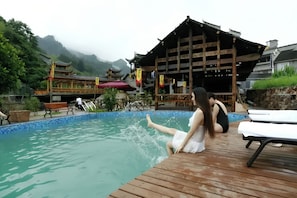 Una terraza de madera con piscina, un edificio tradicional con techo inclinado y montañas al fondo.