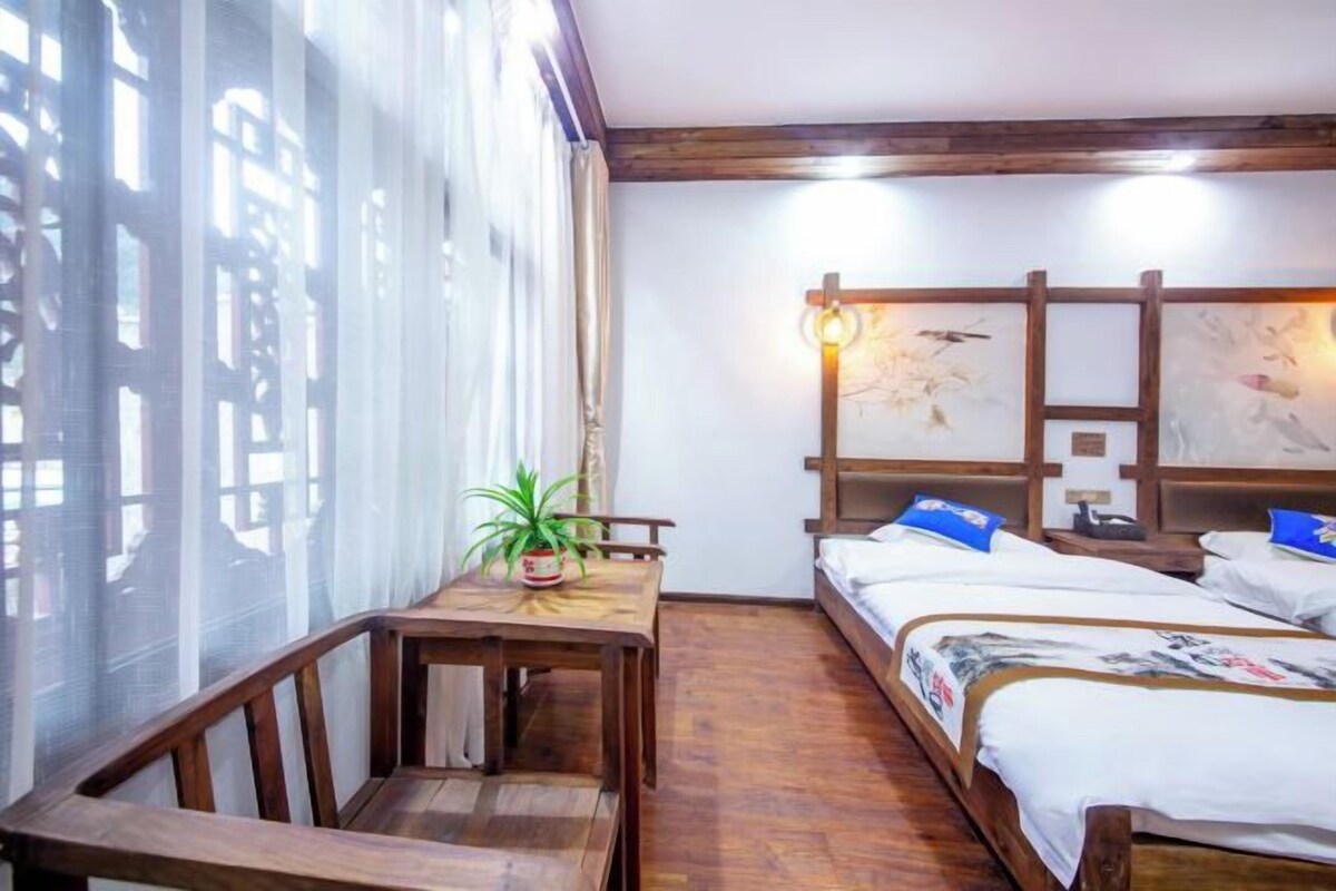 A room with a bed, a table with a plant, wooden beams on the ceiling, and a wall with artwork.