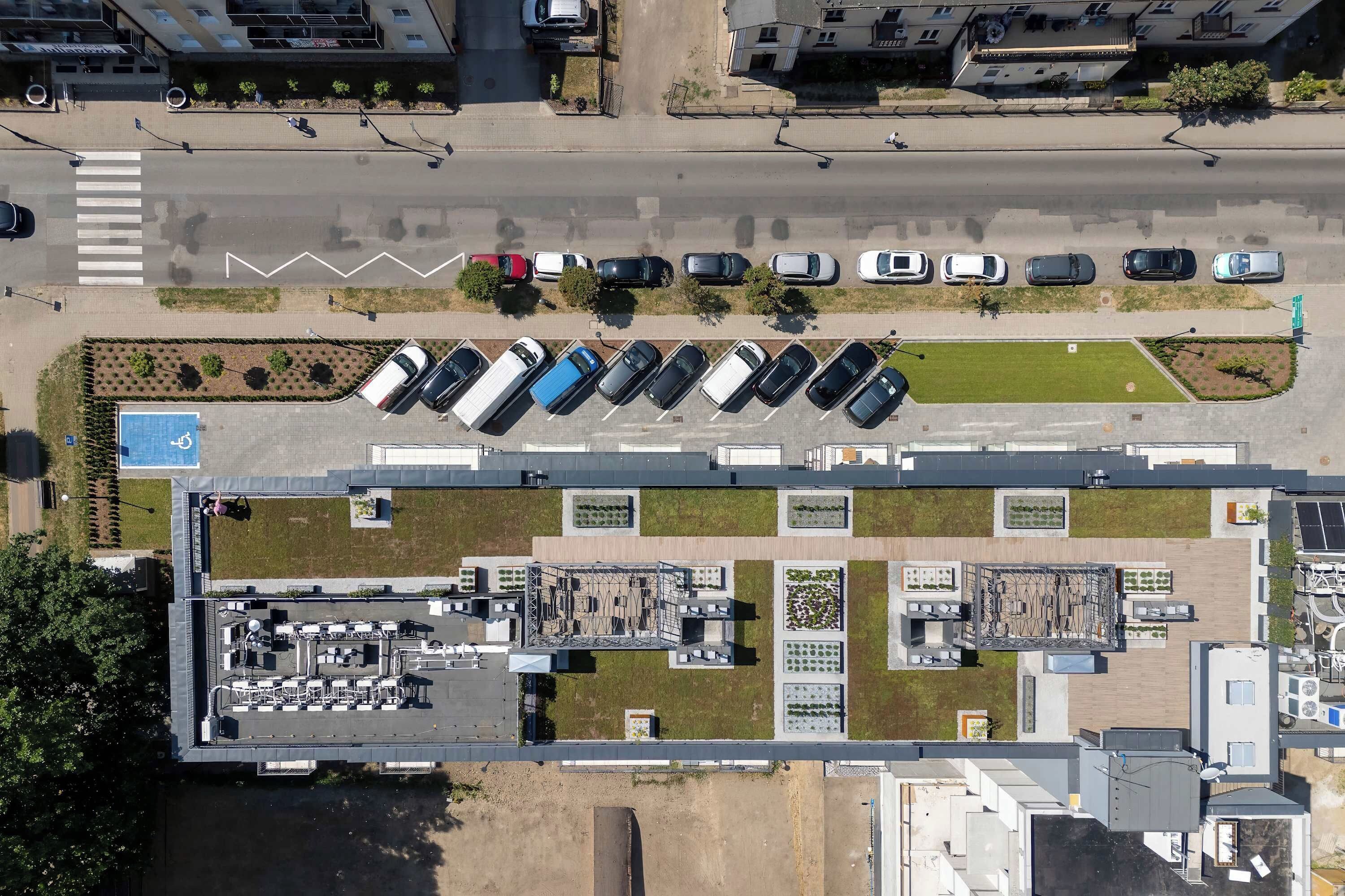 An aerial view of a parking lot with multiple rows of parked cars, a pedestrian crossing, and a building with a flat roof and solar panels.