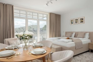 Ein helles Zimmer mit großem Fenster, einem Bett, einem Esstisch mit Stühlen und einer Vase mit Blumen.