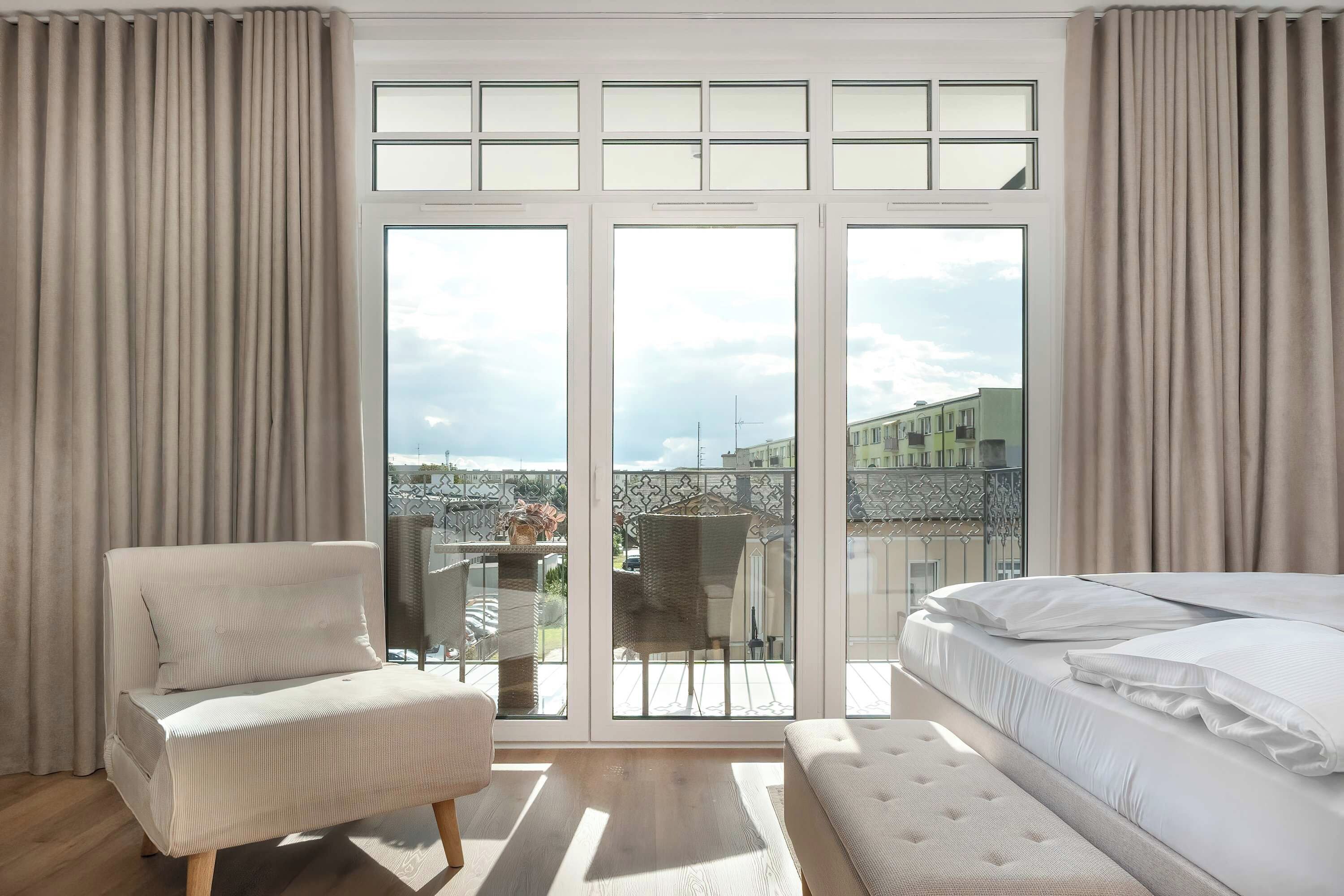 Une chambre dotée d’une grande porte-fenêtre coulissante donnant sur un balcon, avec un lit, une chaise et une vue sur les immeubles.