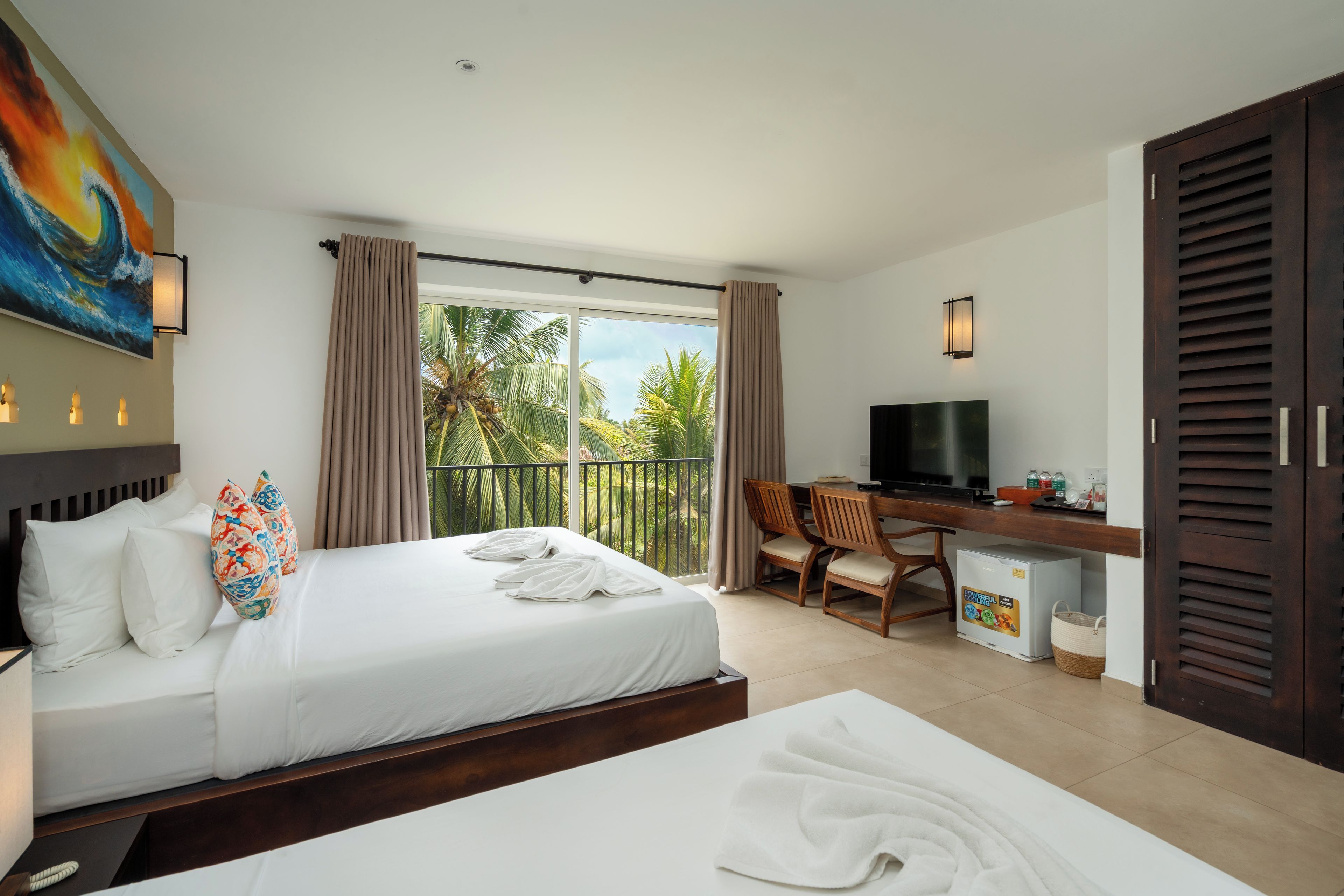 Habitación de hotel con cama, escritorio, televisor y balcón con vista a palmeras.