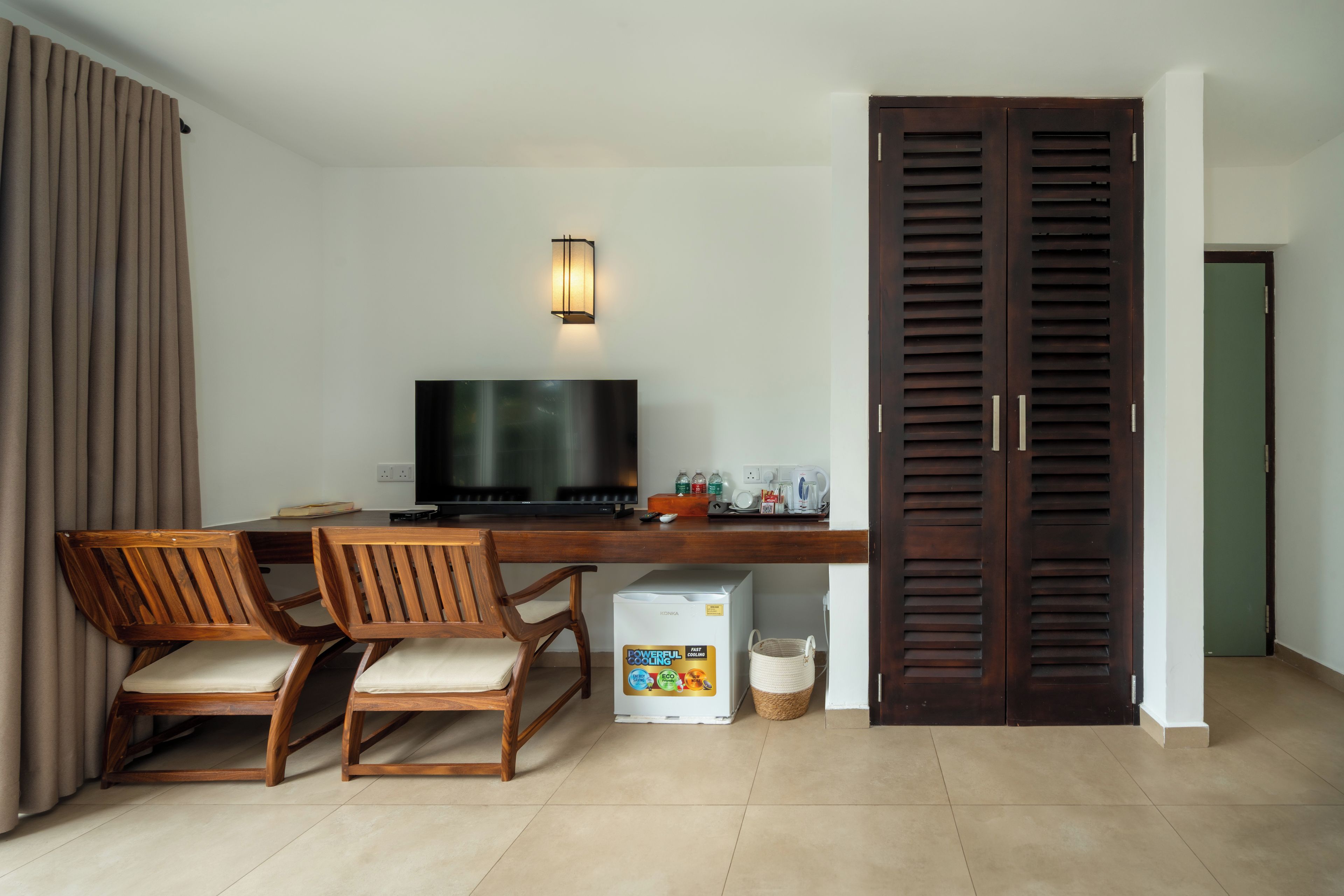 Ein Zimmer mit Schreibtisch, zwei Stühlen, einem Fernseher, einem Kleiderschrank und einem kleinen Kühlschrank.