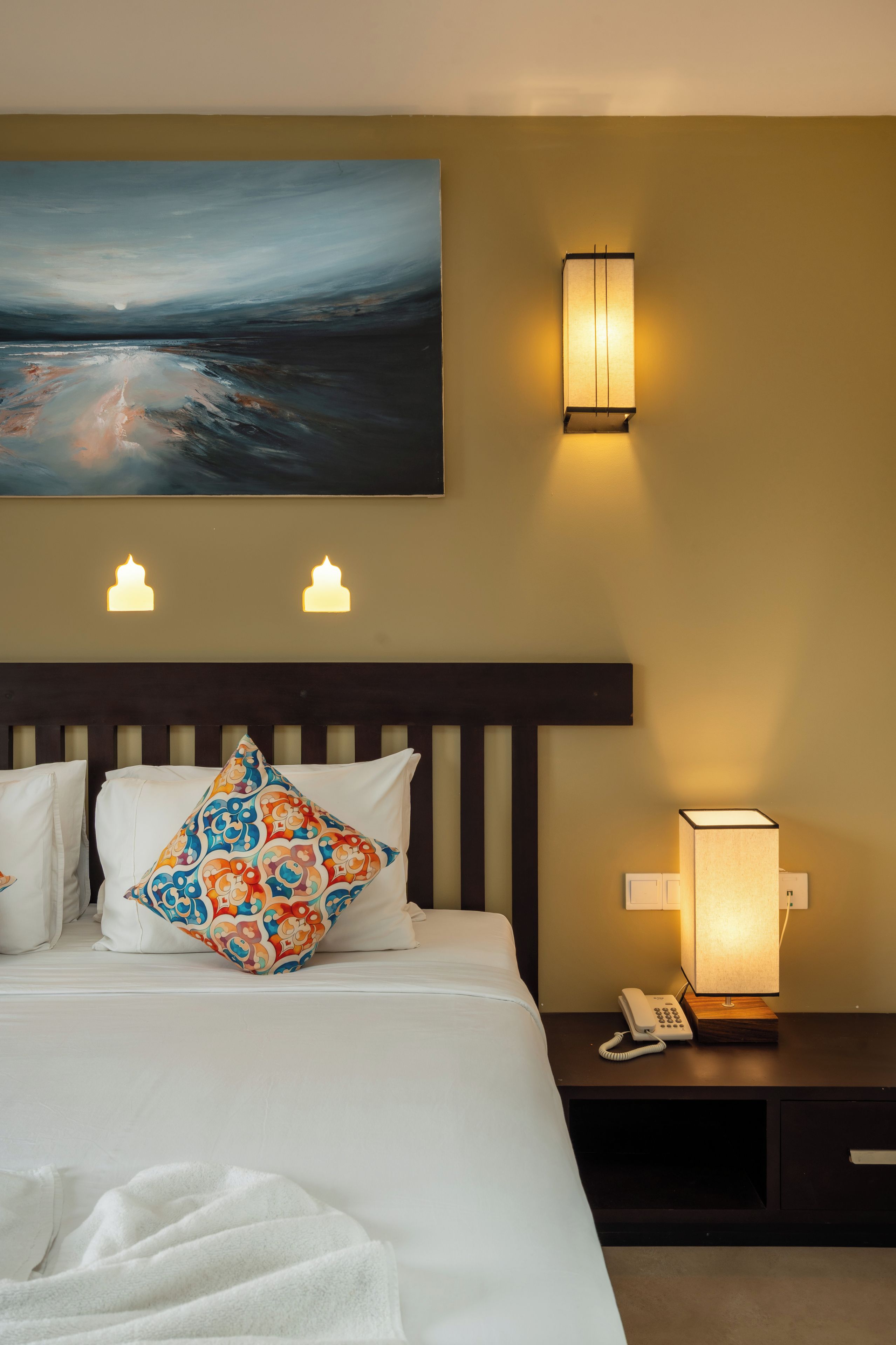 Habitación de hotel con cama, mesita de noche con lámpara, teléfono y un cuadro en la pared.