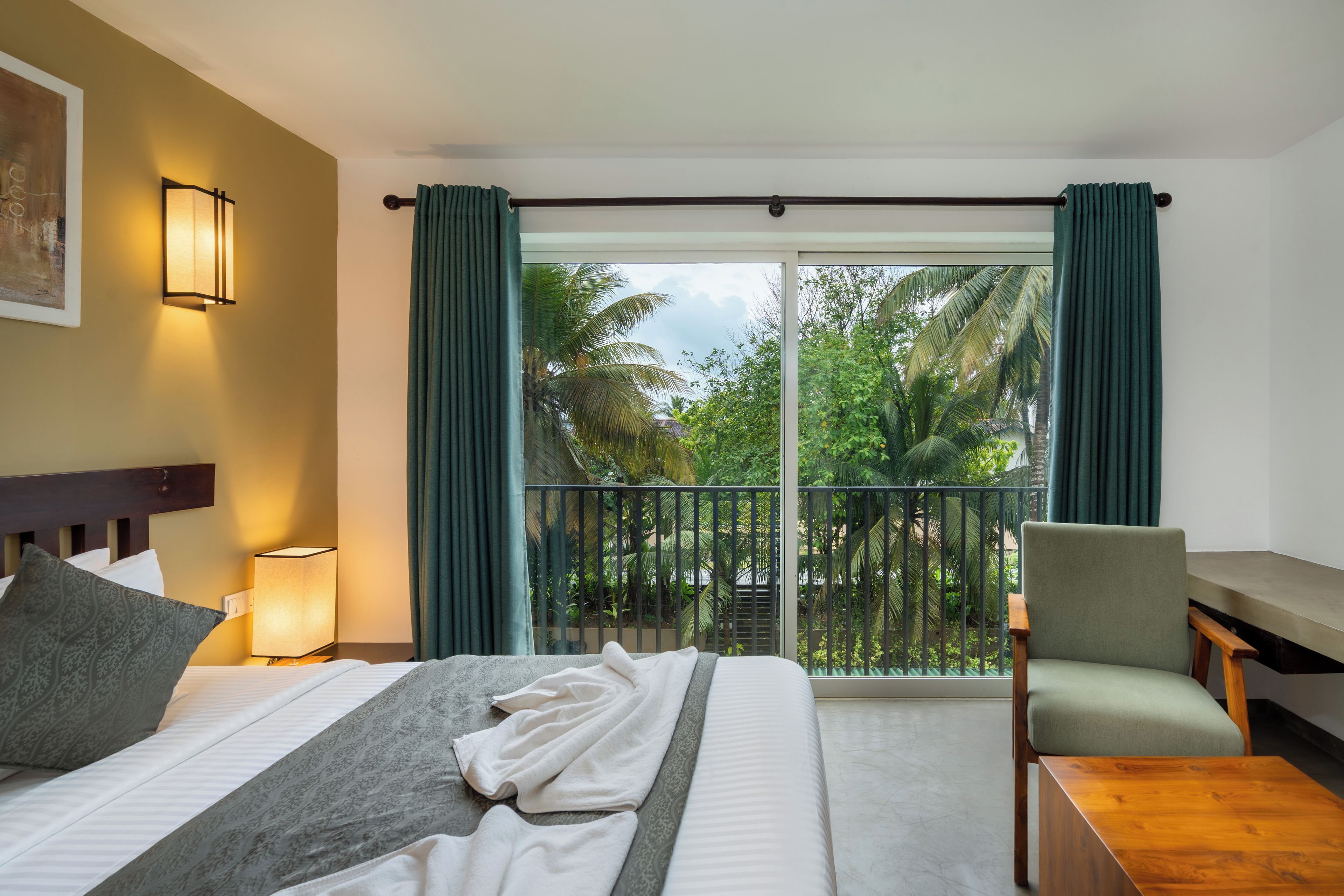 A hotel room with a bed, a chair, a table, a nightstand with a lamp, and a view of palm trees through a glass door.