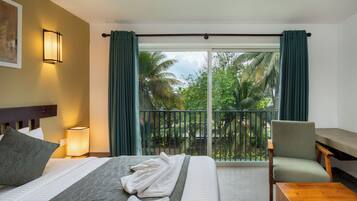 A hotel room with a bed, a chair, a table, a nightstand with a lamp, and a view of palm trees through a glass door.