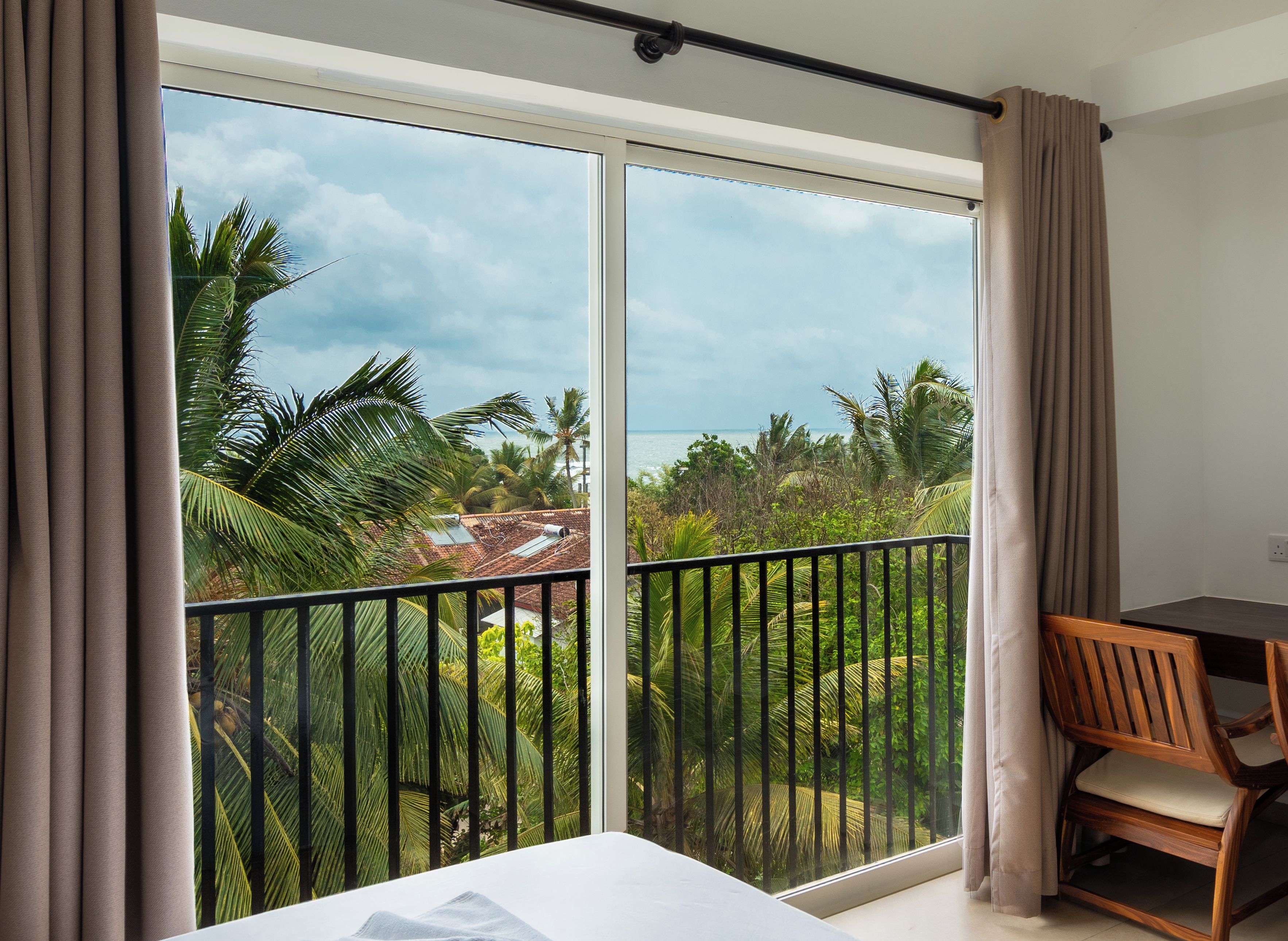 A room with a bed, a desk, and a view of a tropical landscape through a large window.