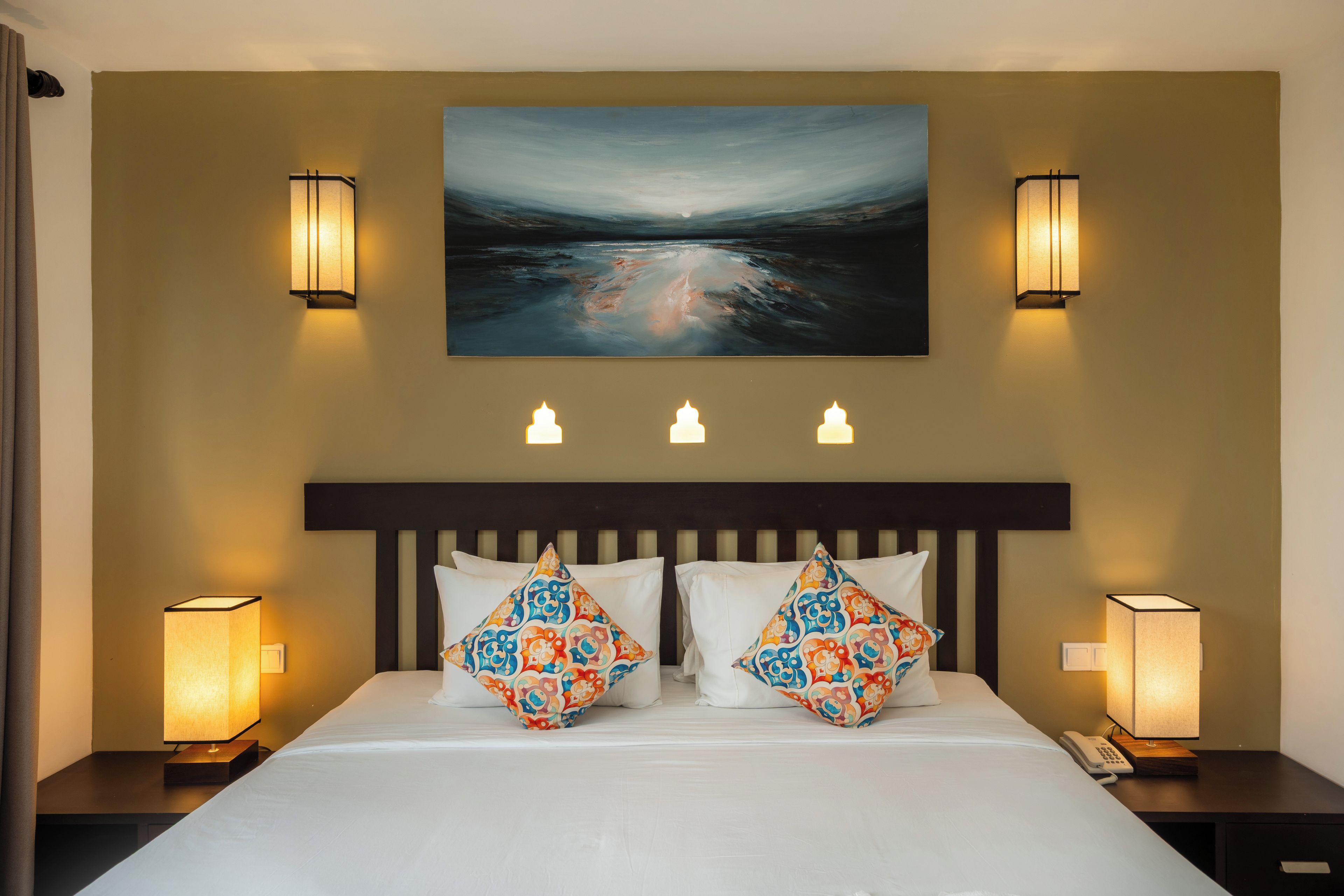 A hotel room with a large bed, two bedside lamps, a painting above the bed, and a telephone on the nightstand.