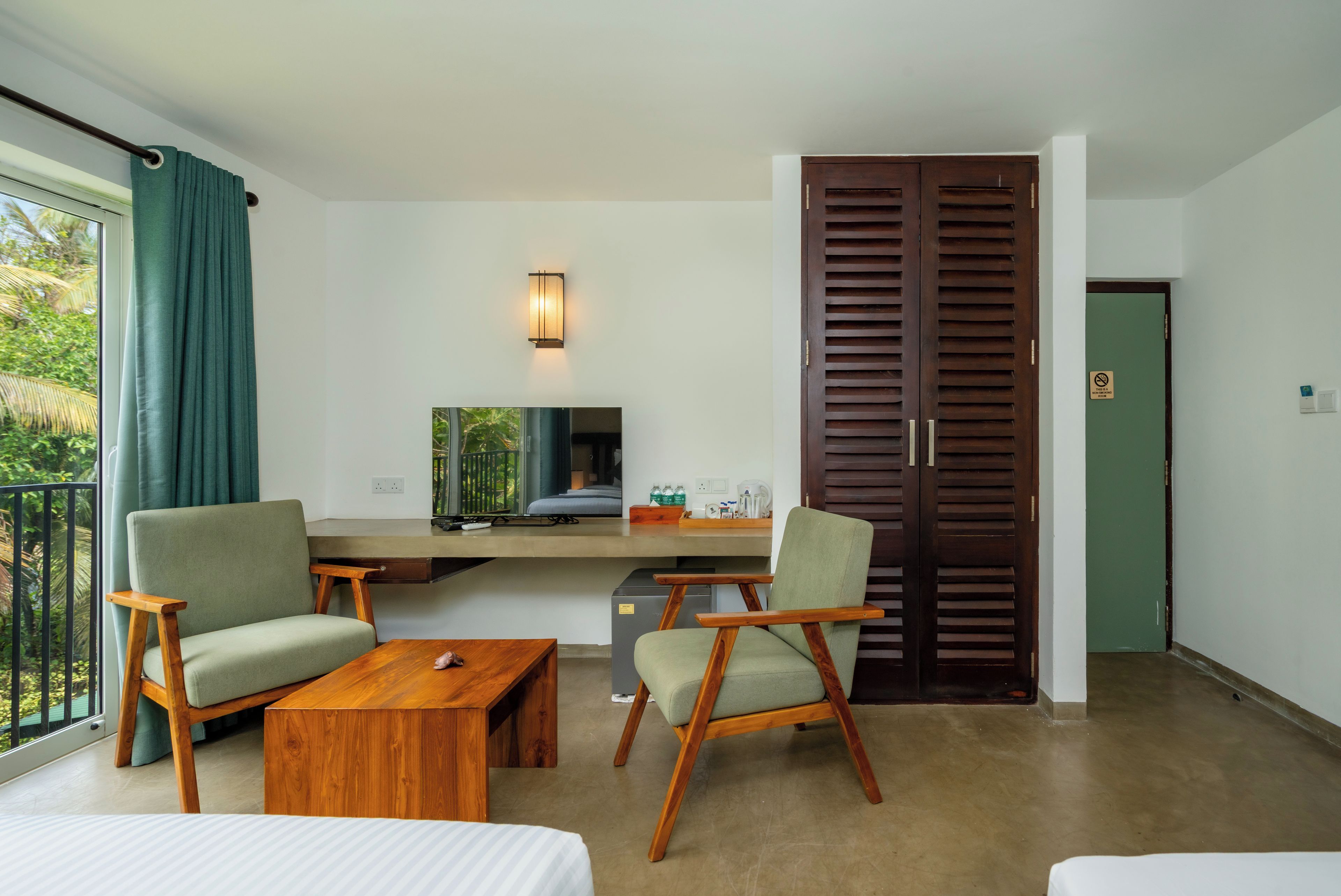 A hotel room with a bed, two armchairs, a wooden coffee table, a desk with a computer, a TV, and a large wooden door.