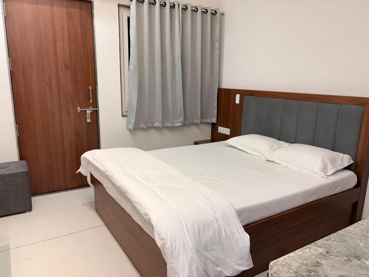 Un dormitorio con cama de madera, cortinas grises, una puerta de madera y un banco gris.