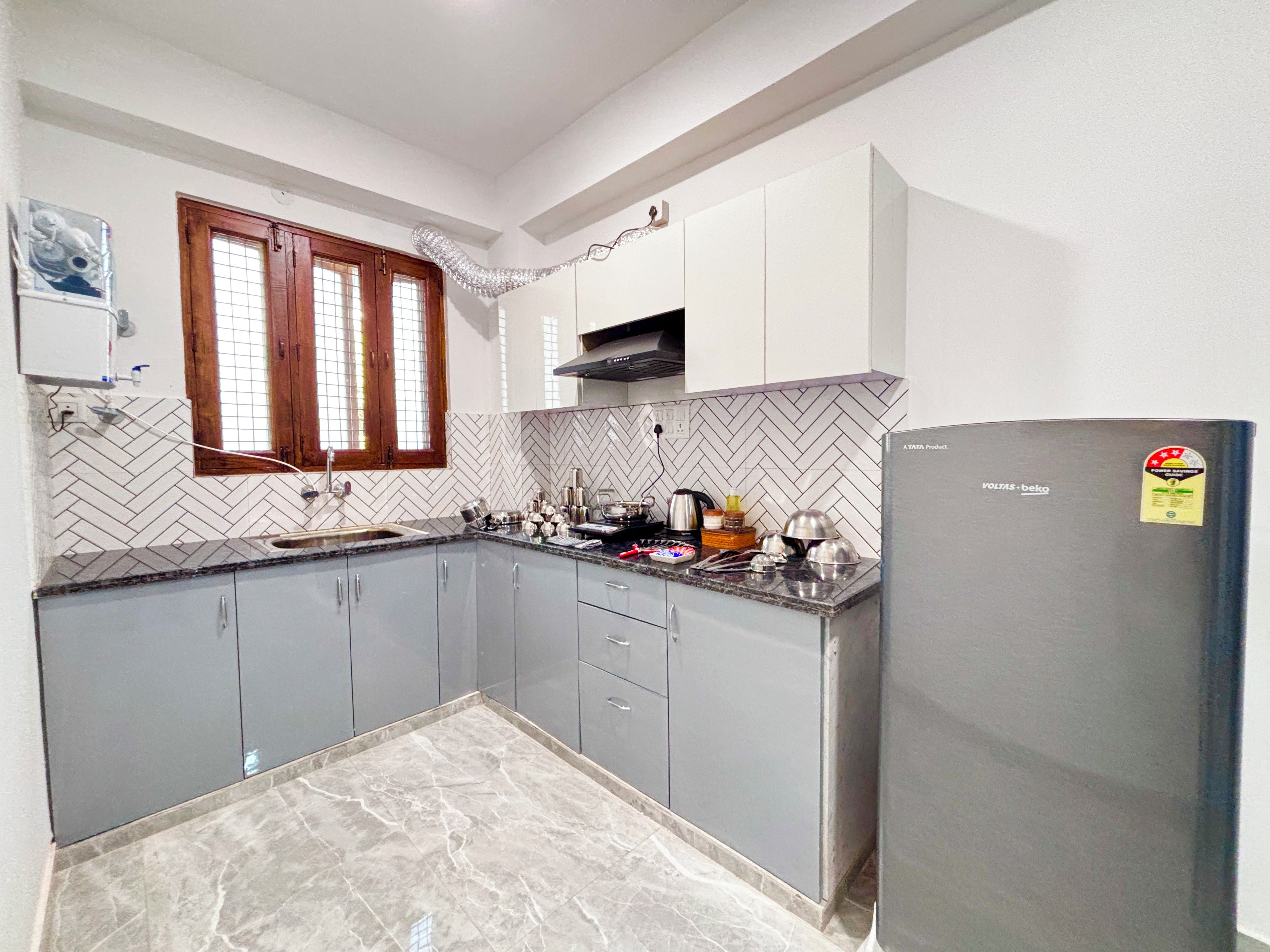 A kitchen with grey cabinets, a herringbone tile backsplash, a window with wooden frames, and a refrigerator with a sticker.