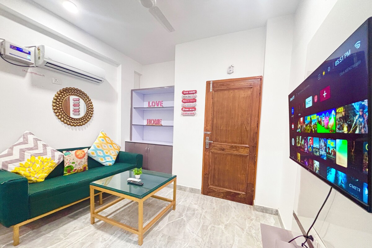 A living room with a green sofa, a glass coffee table, a TV mounted on the wall, and a wooden door.