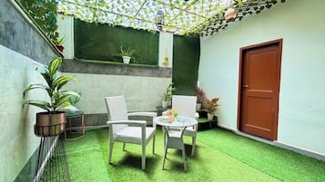 A small patio area with a green artificial turf floor, two wicker chairs, a small round table, and potted plants.