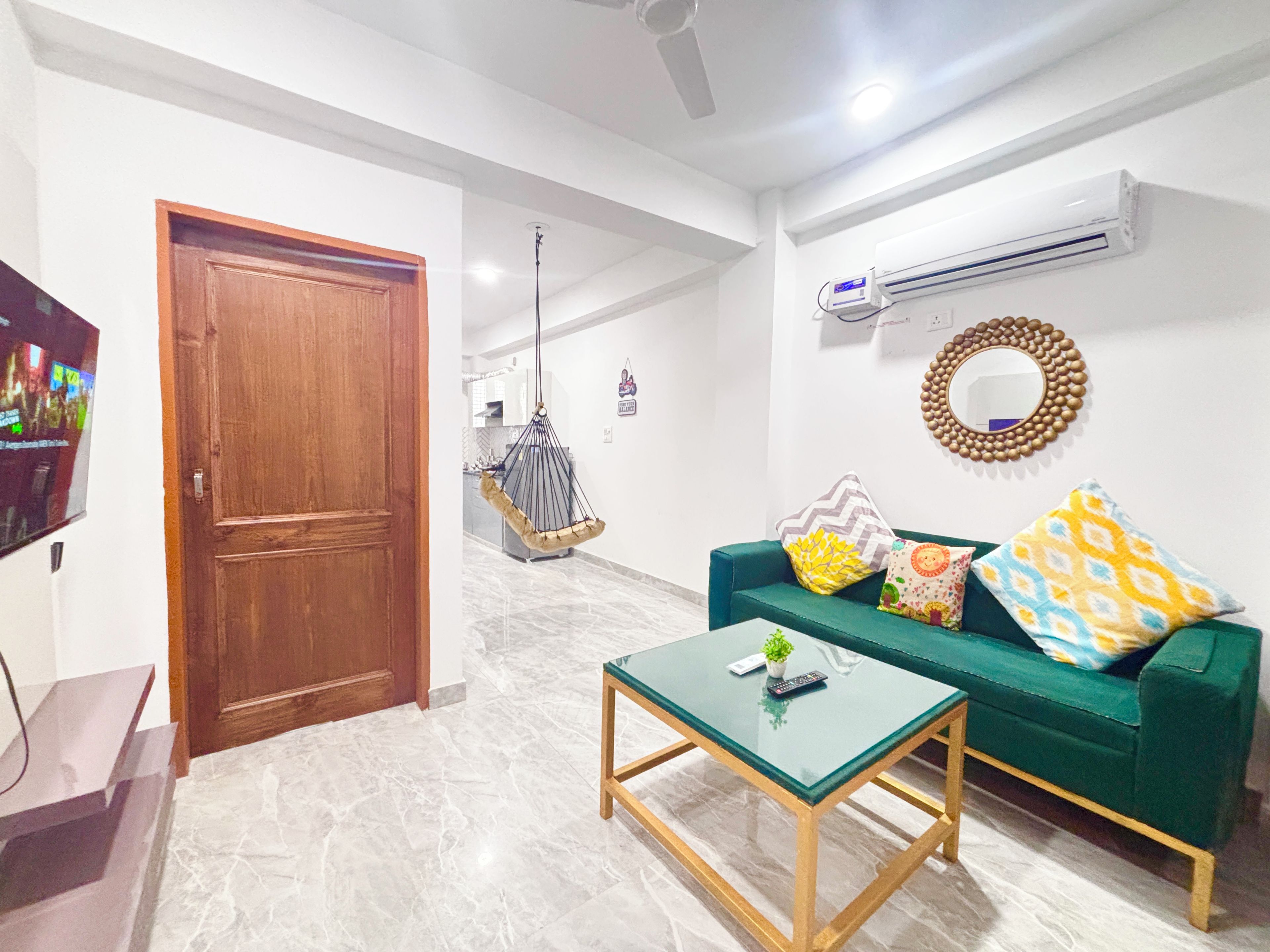 Una sala de estar con un sofá verde, una mesa de centro de vidrio, una puerta de madera y un televisor a la izquierda.