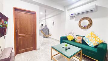 Una sala de estar con un sofá verde, una mesa de centro de vidrio, una puerta de madera y un televisor a la izquierda.