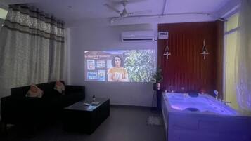 A living room with a black sofa, a coffee table, a large screen, and a bathtub with a blue light.