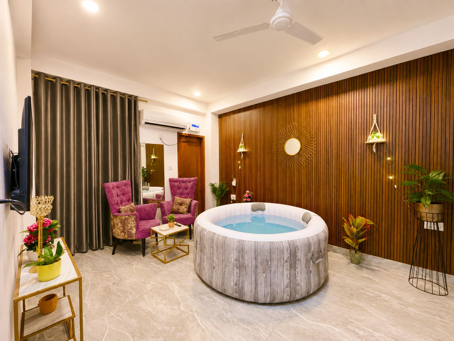 Una sala de estar moderna con una bañera de hidromasaje circular, dos sillones morados, una mesita y un televisor montado en la pared.