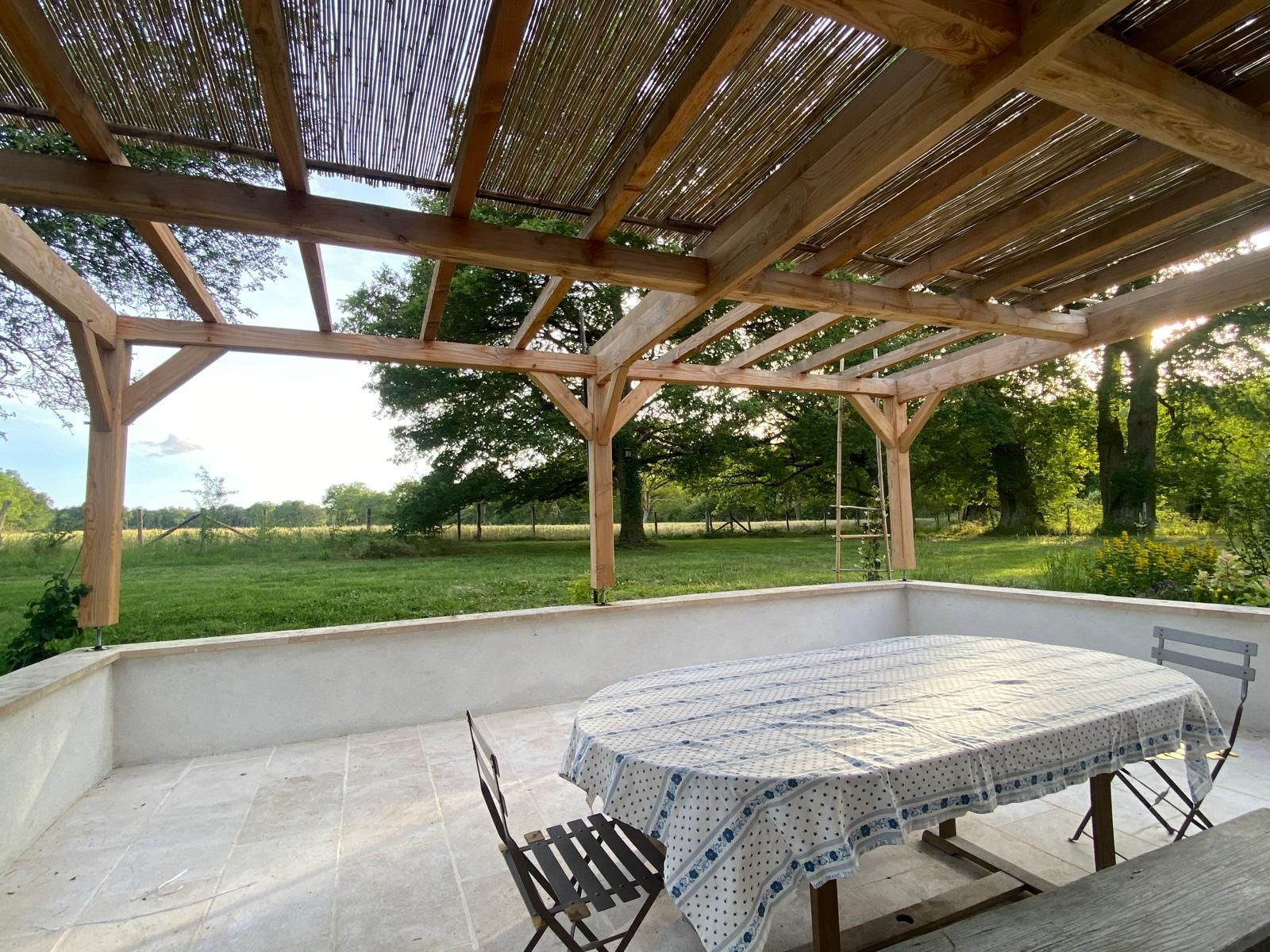 A covered outdoor patio with a table and chairs, surrounded by a wooden pergola and a view of a grassy field.