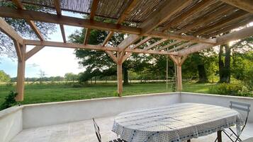 A covered outdoor patio with a table and chairs, surrounded by a wooden pergola and a view of a grassy field.