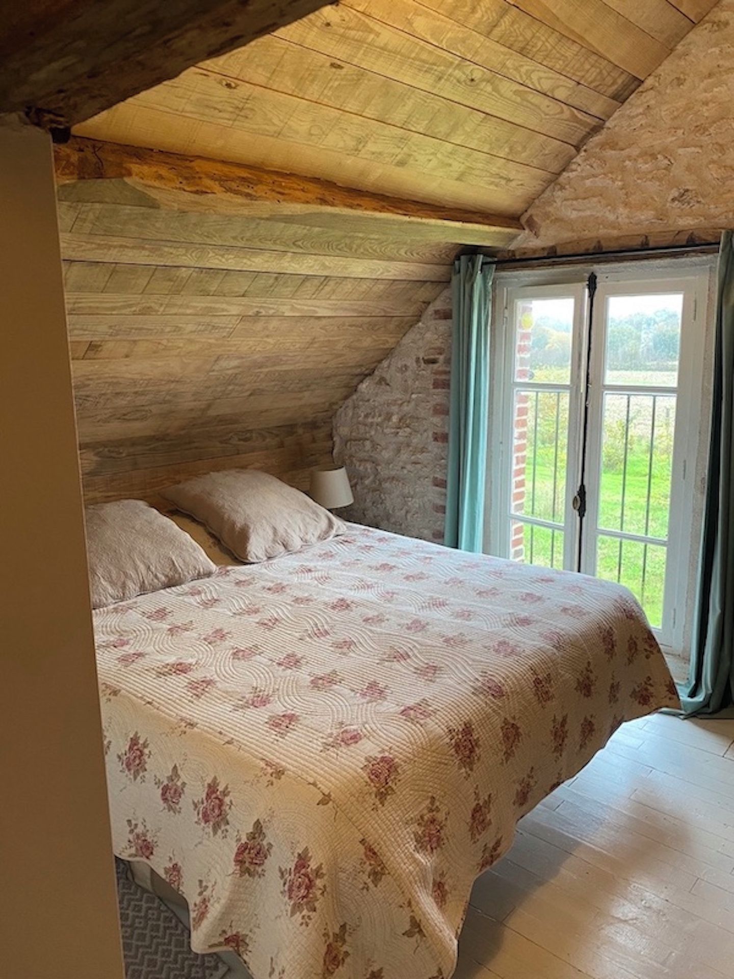 Een slaapkamer met een schuine kap, een bed met bloemenbedekking, een nachtkastje en een raam met uitzicht op groen.
