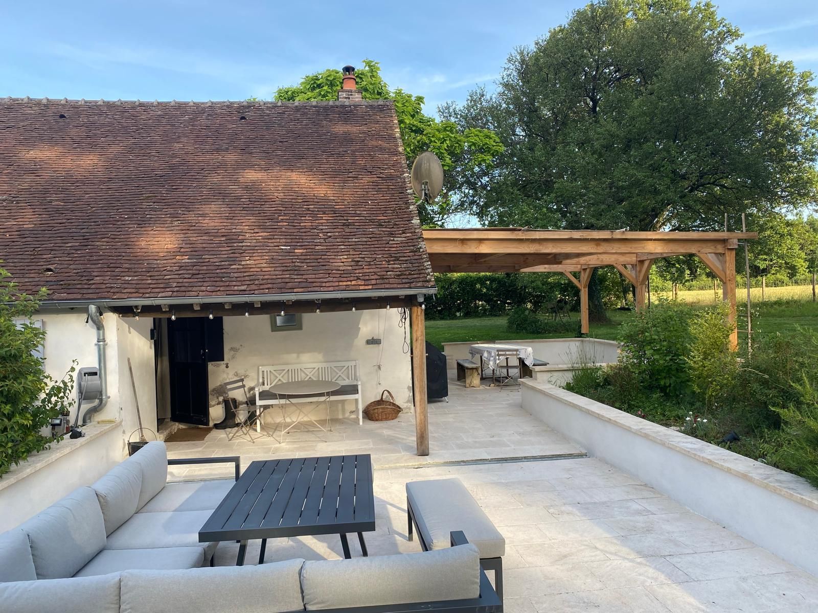 Een rustiek buitenterras met een witte tafel, een bank en een houten pergola.