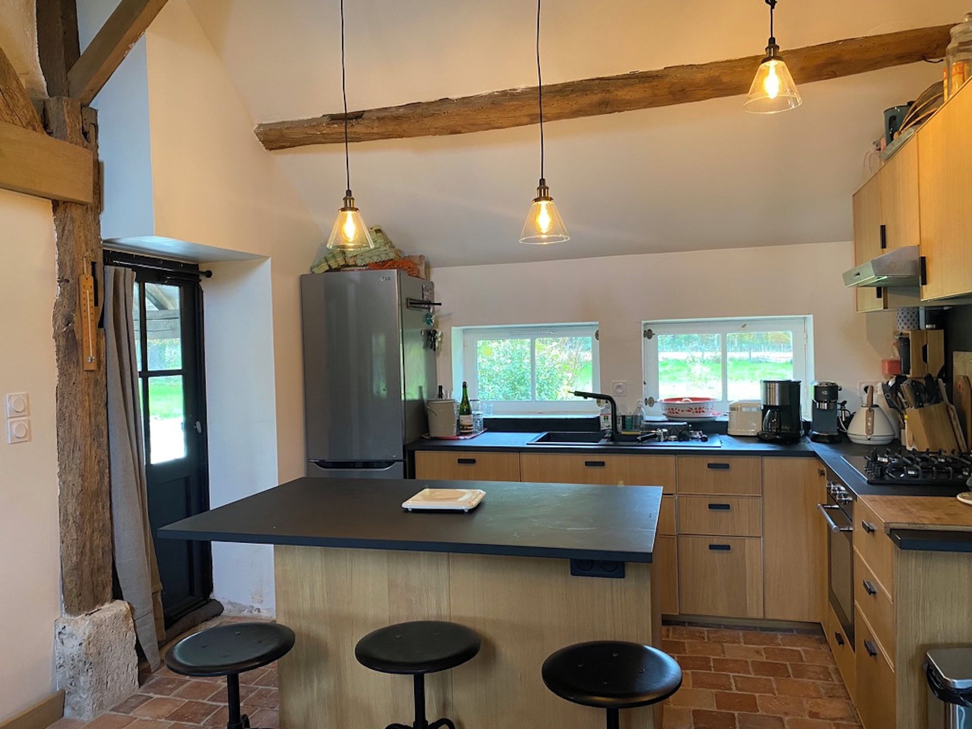 A kitchen with a central island, bar stools, hanging pendant lights, and a view of the outdoors through windows.