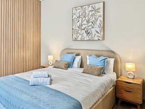 Ein ordentlich gemachtes Bett mit einer blauen, gemusterten Decke, zwei Nachttischlampen und einem Wandbild darüber.
