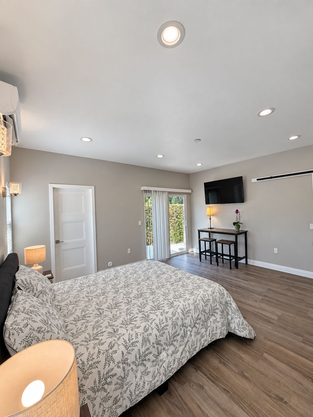 A bedroom with a bed, bedside lamps, a desk with a chair, a television, and a sliding glass door leading to a balcony.